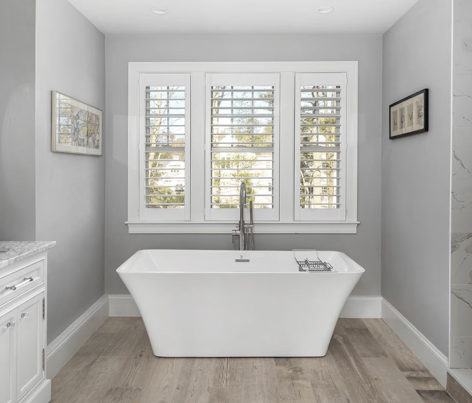 Modern gray bathroom with a freestanding tub beneath three windows and white cabinetry on the left.