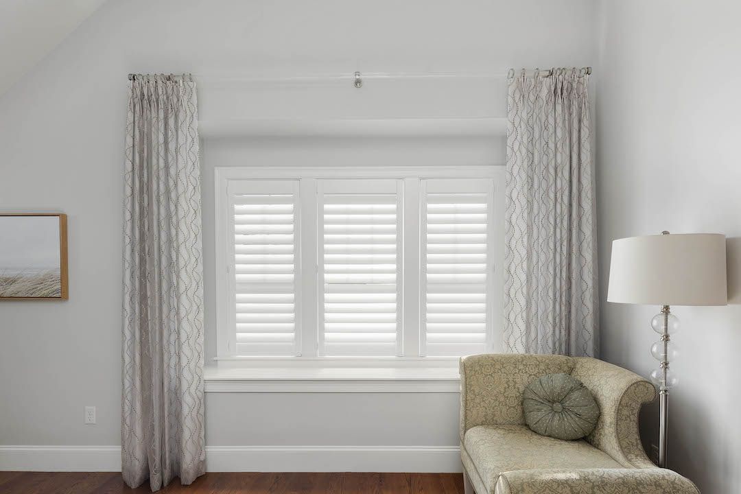 Bright living room with white shutters, patterned curtains, a beige chair, and a floor lamp on a hardwood floor