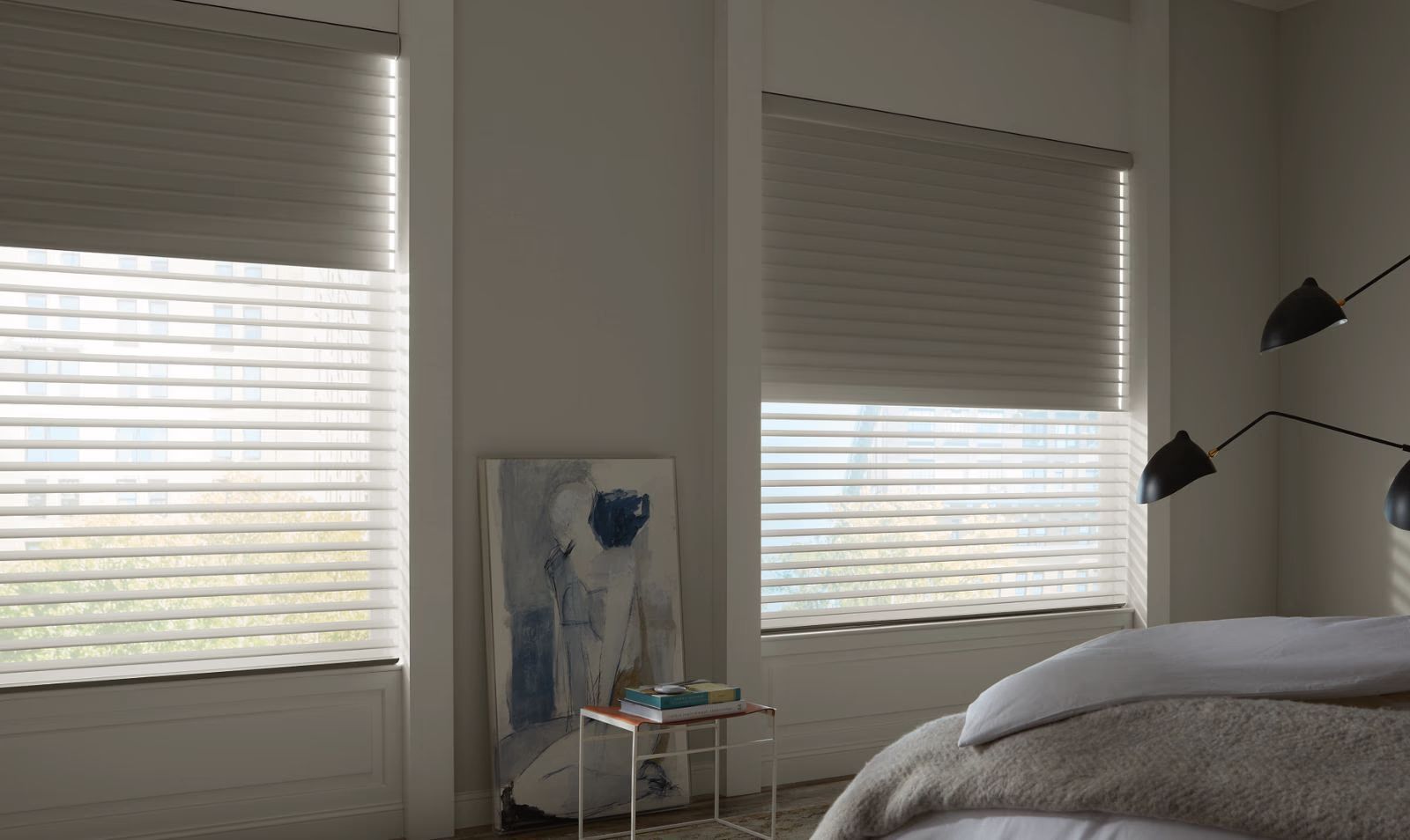 Bright bedroom with two window blinds, a bedside lamp, and a bed in the foreground.