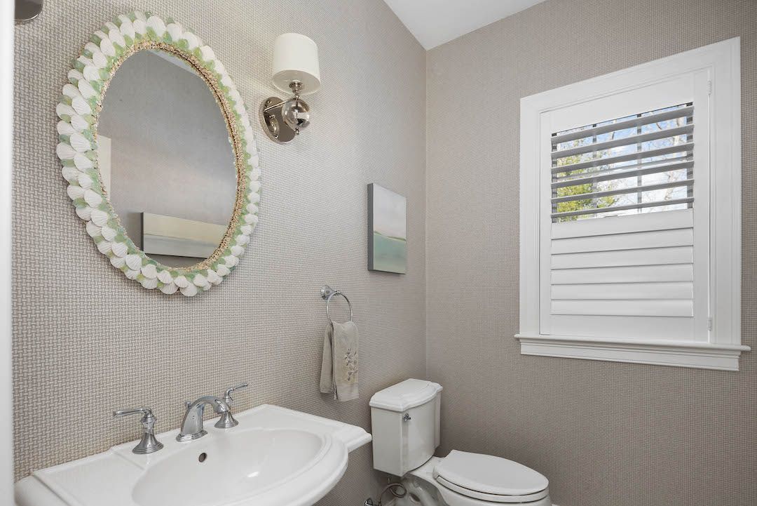 Small beige bathroom with white sink, toilet, oval mirror, and window shutters.