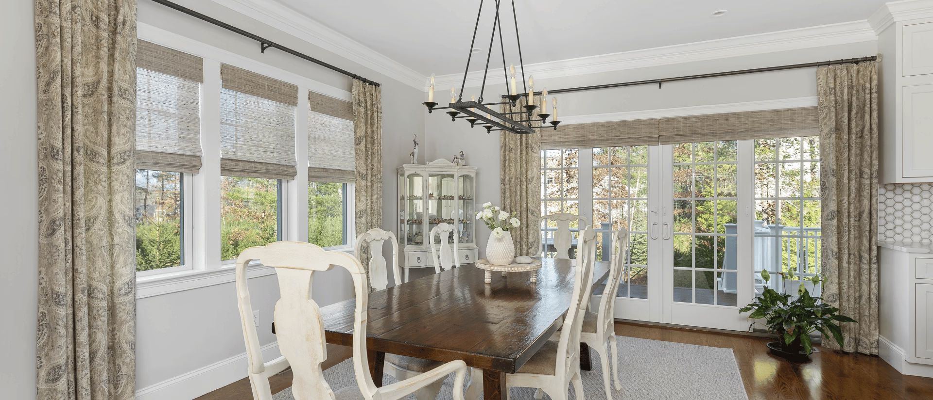 Bright dining room with a dark wood table, white chairs, large windows, and patterned curtains