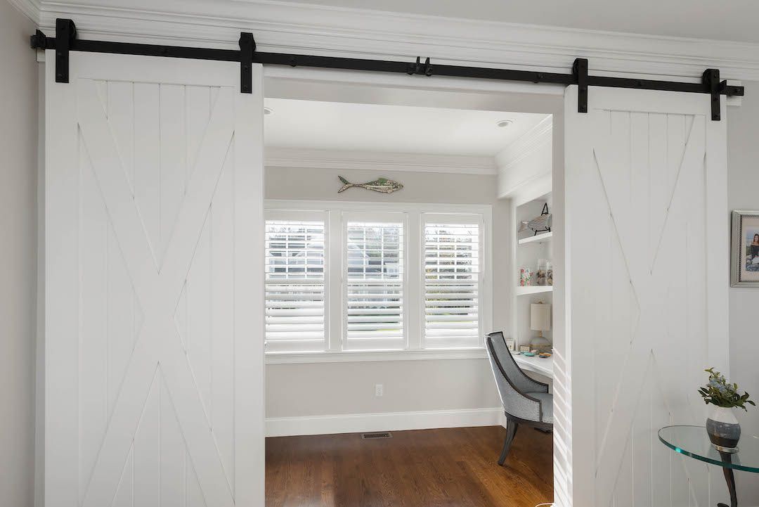 White sliding barn doors open to a bright room with windows, hardwood floor, and a chair by the desk.