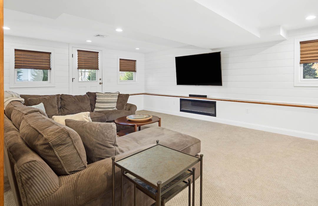 Bright basement living room with beige couches, wall-mounted TV, small windows, and carpeted floor
