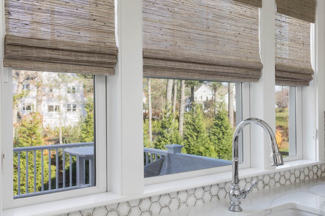 Kitchen sink with chrome faucet beneath woven shades, overlooking a patio and greenery.