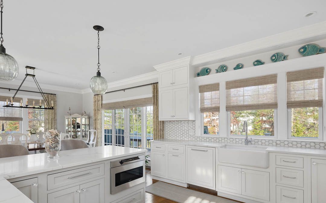 Bright white kitchen with large windows, pendant lights, and decorative cabinets beneath a sunny view.