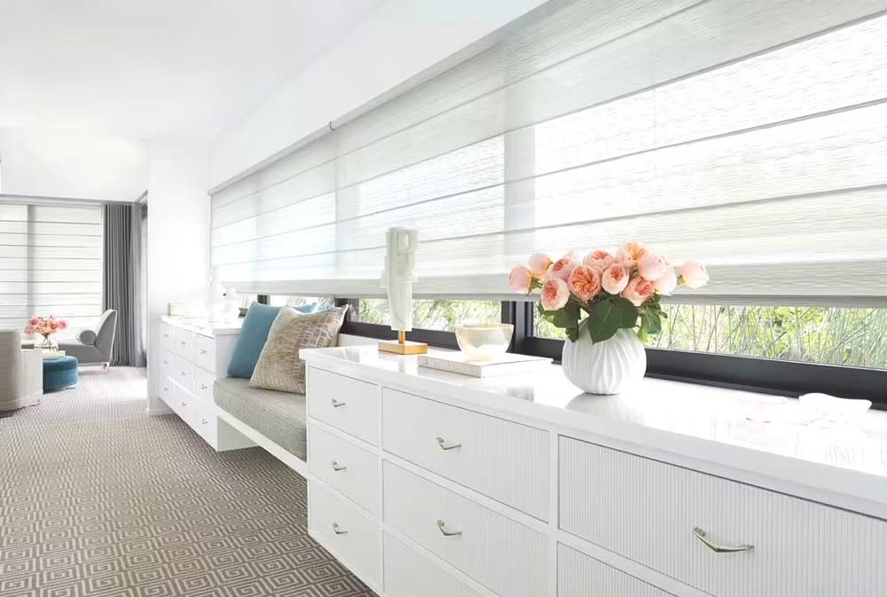 Bright white bedroom with bed, dresser, gray carpet, and flowers by large windows