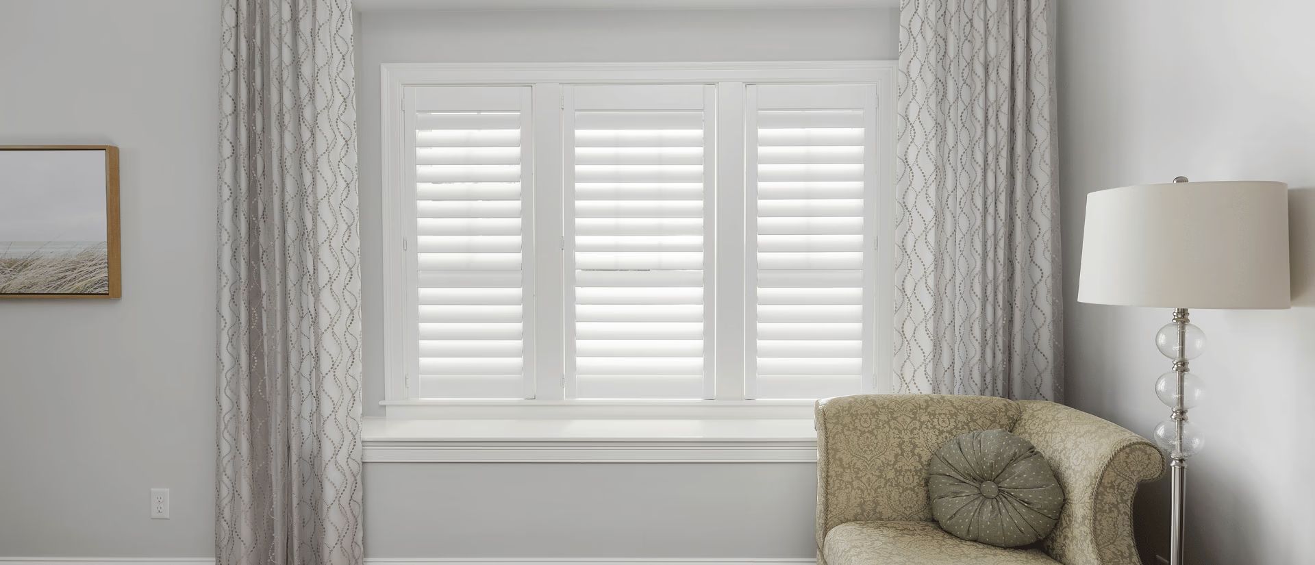 Bright living room corner with white shutters, patterned curtains, a beige chair, and a floor lamp.