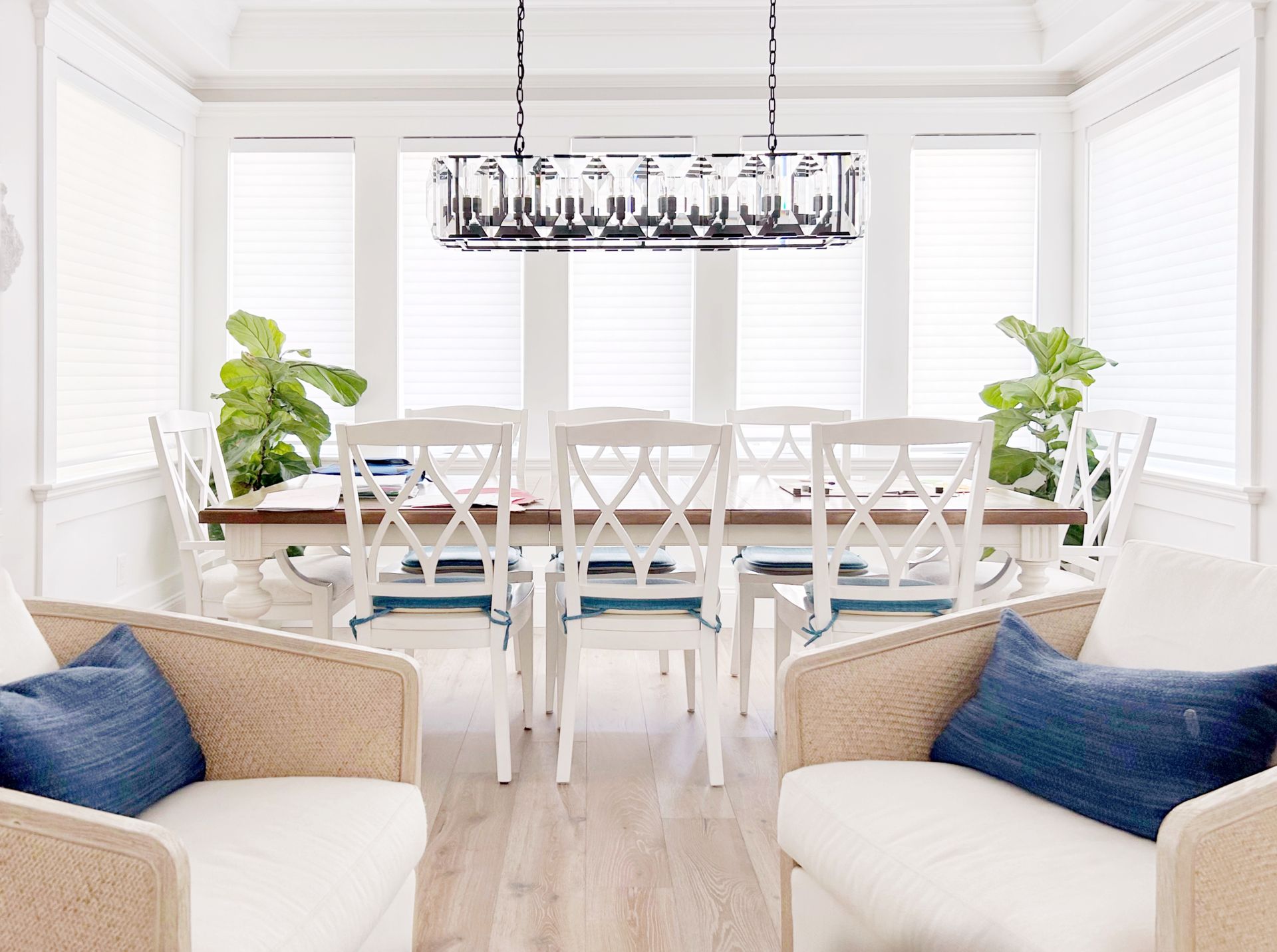 Bright dining room with a long white table, chandelier, blue chairs, and large windows, seen from a living area