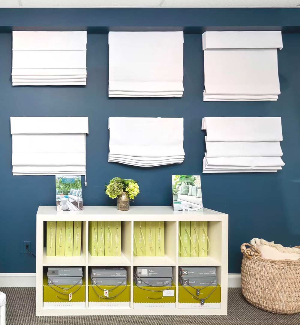 Neatly organized blue wall with white window blinds and a white cubby shelf holding books, plants, and baskets
