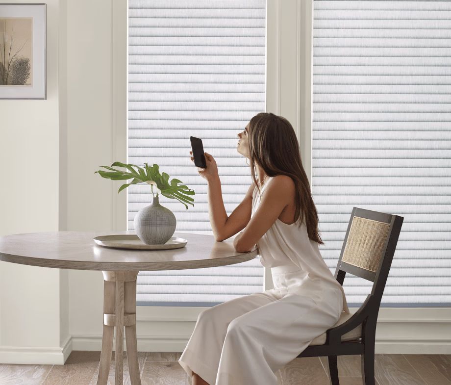Woman in white dress using a phone at a round table beside a potted plant, near bright window blinds