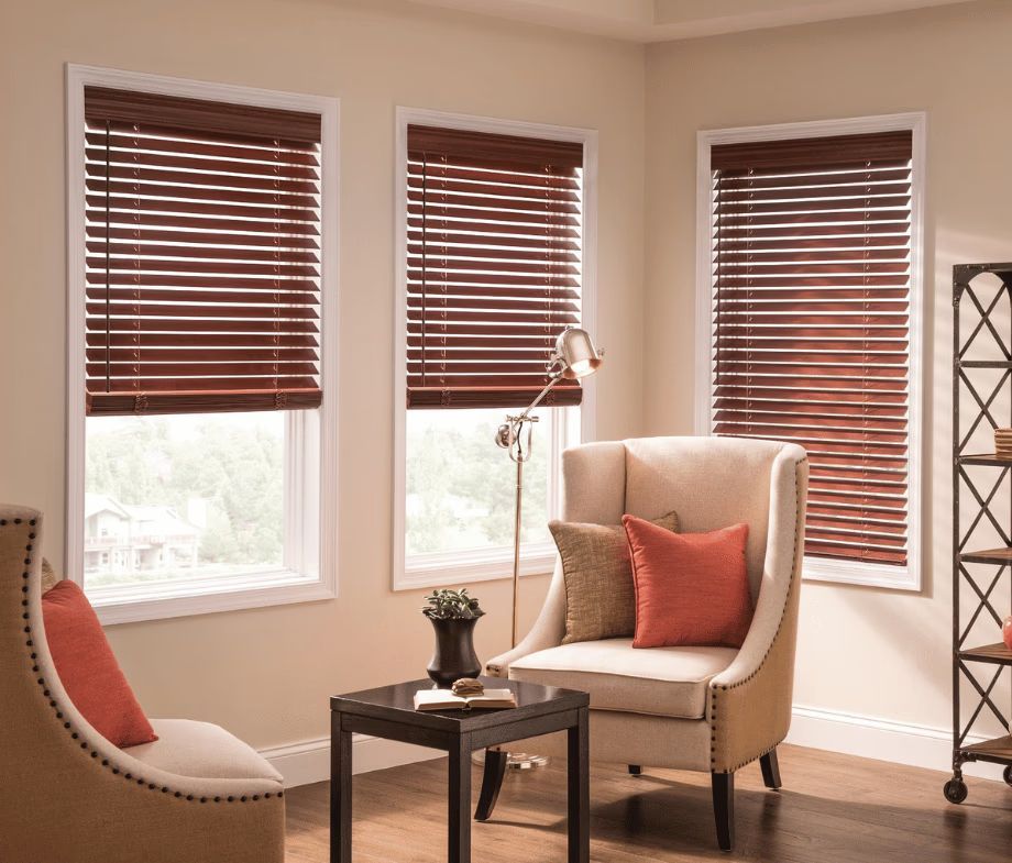 Cozy living room with three windows, brown blinds, beige chairs, and a small table by sunlight