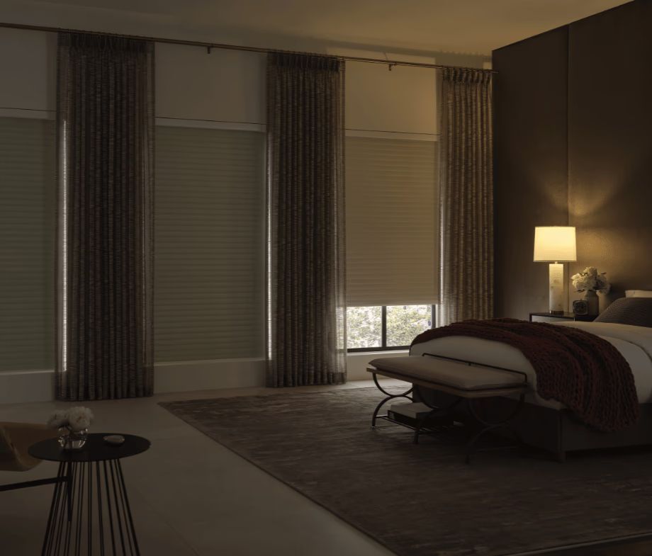 Dim bedroom with a made bed, floor lamp, curtains, and a chair by the window.