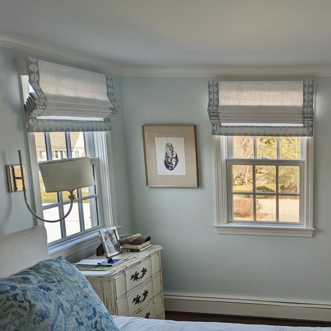 Bright bedroom corner with blue bedspread, framed art, window blinds, and a small white dresser.
