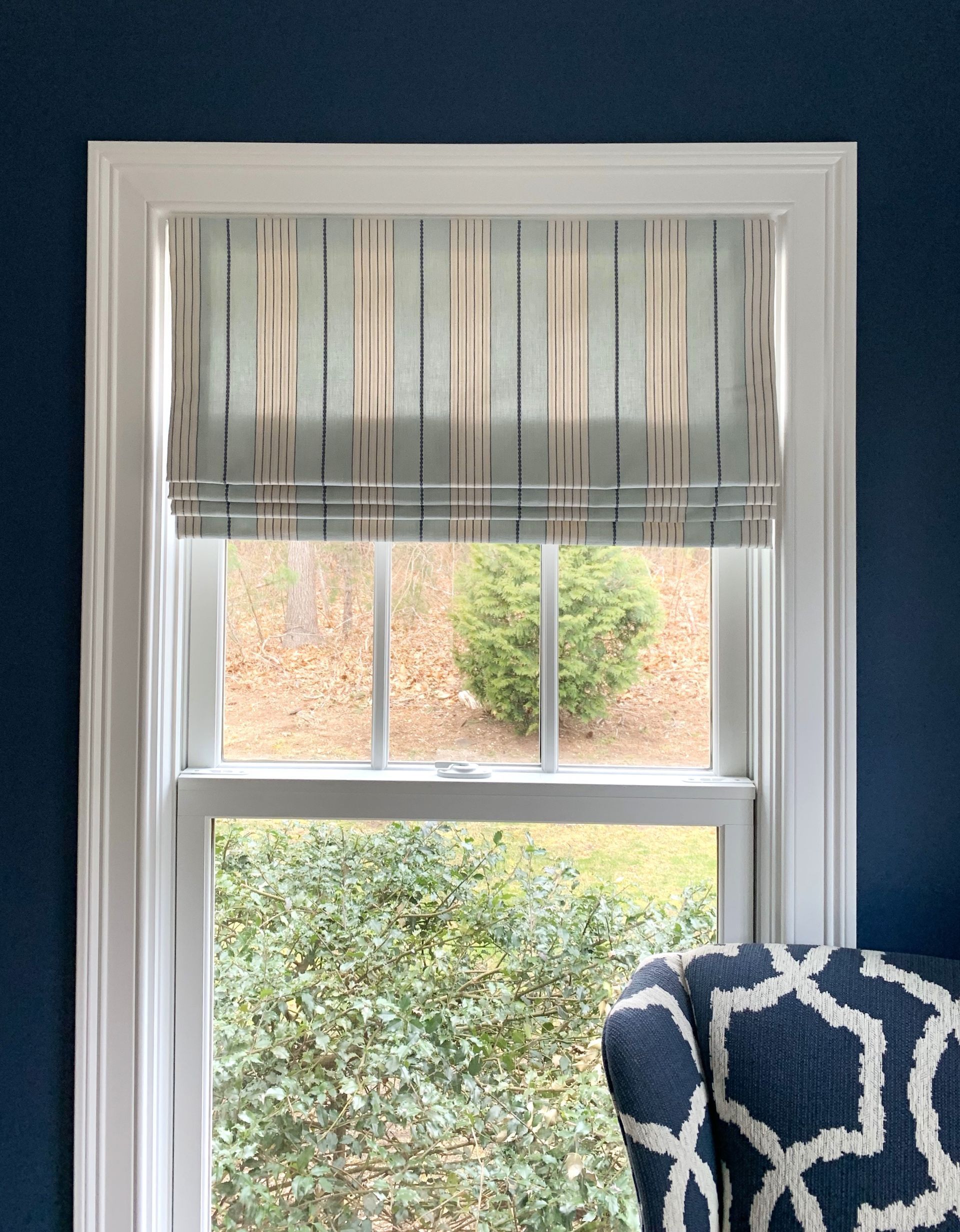 Window with striped roman shade, white trim, and a blue-and-white patterned chair corner.