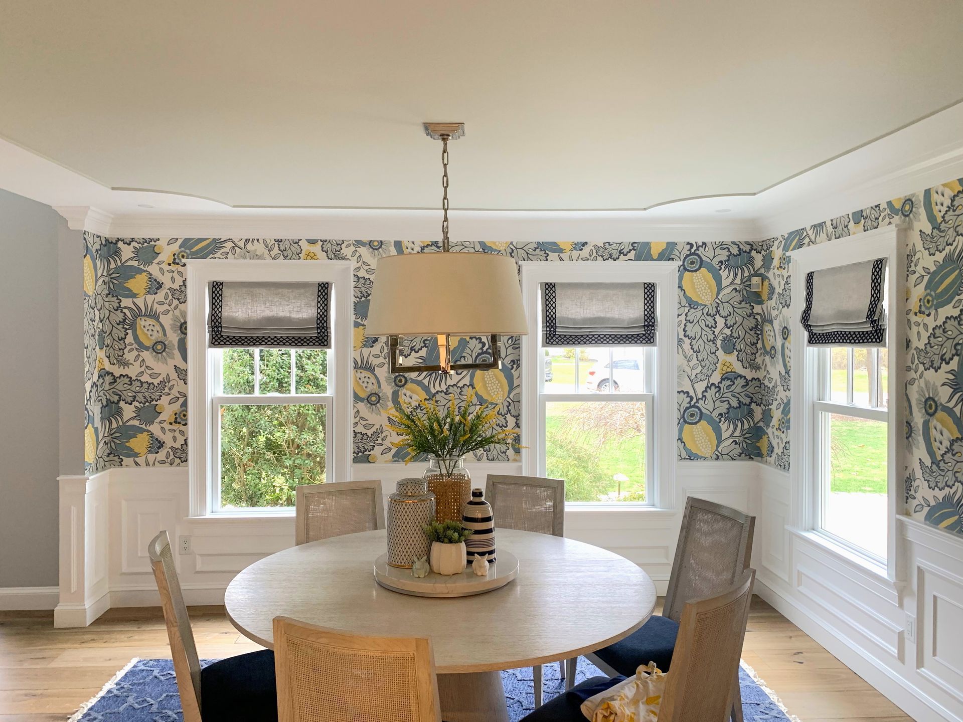 Bright dining room with floral wallpaper, round wooden table, upholstered chairs, and three windows with blinds