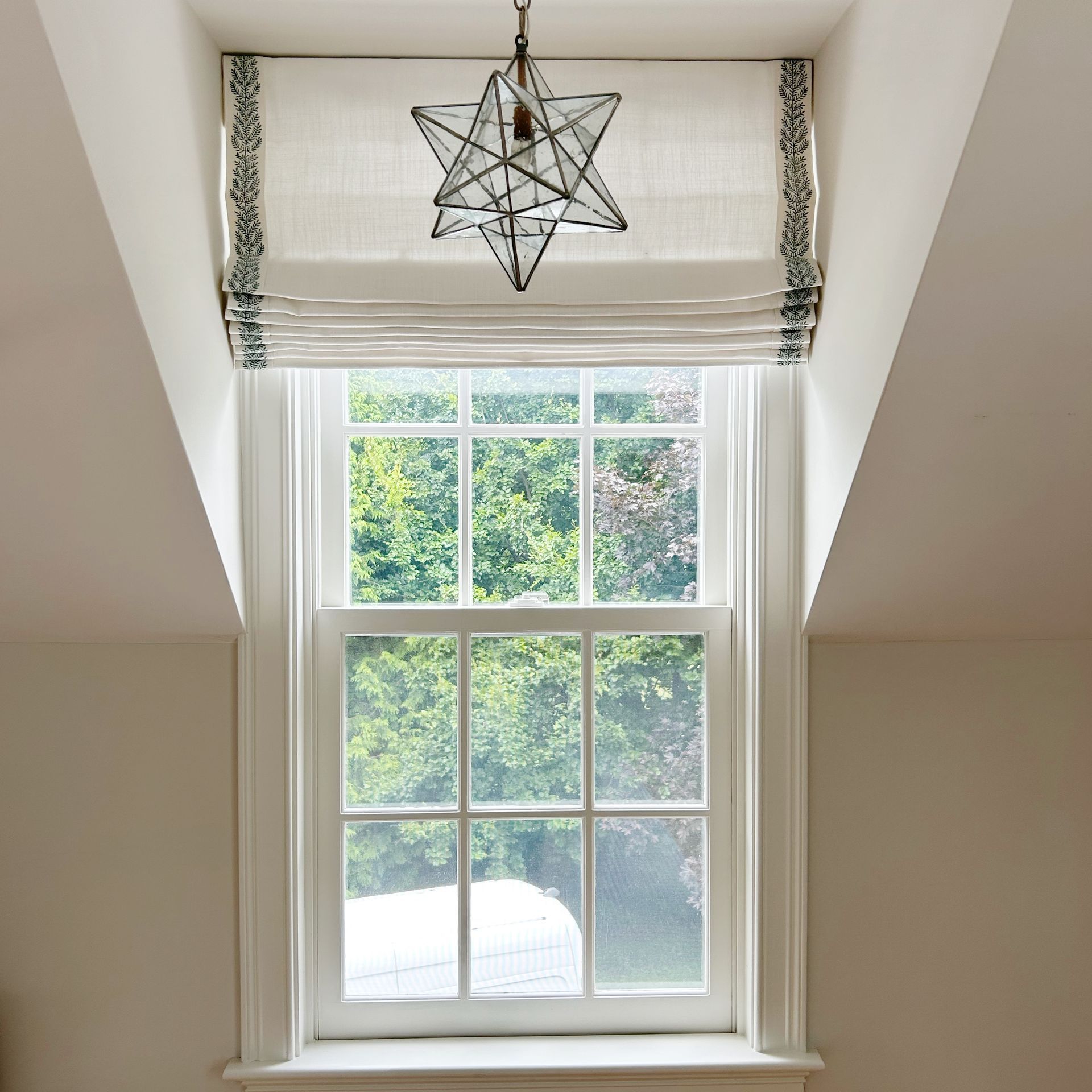Bright window in a sloped white room with a hanging star lantern and green trees outside.