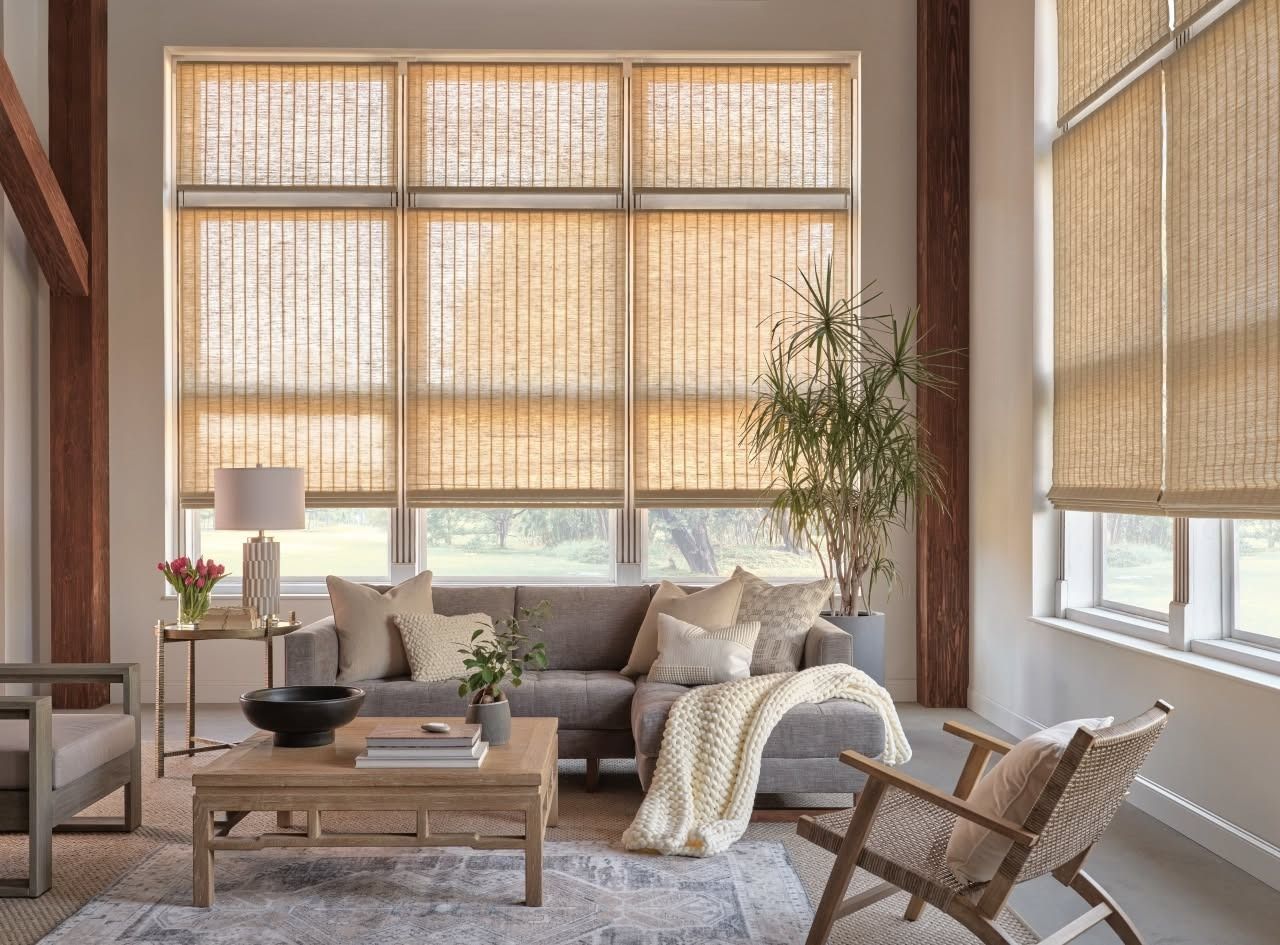 Bright living room with woven blinds, neutral sofa, wooden table, chair, and potted plant by large windows