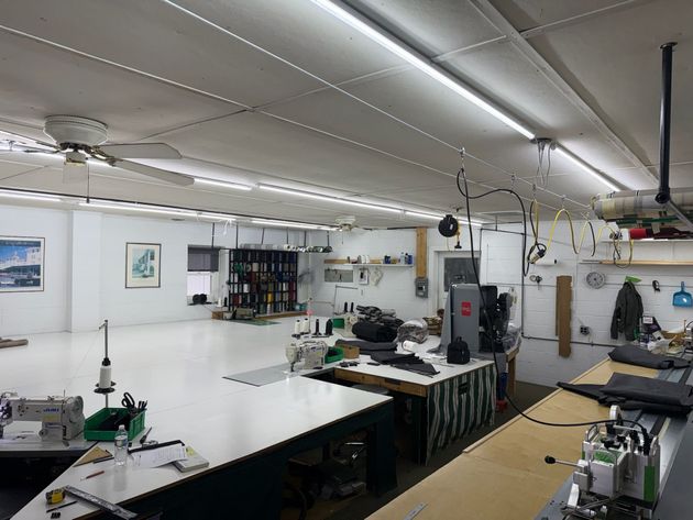 Interior of a sewing workshop with tables, machines, and various supplies; white walls, ceiling, and floor.