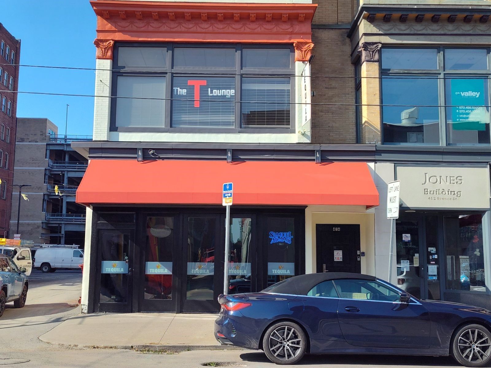 The T Lounge storefront with a bright red awning and upper-floor windows, located on a street with a parked blue car.