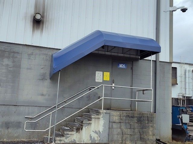 A gray exterior door with a blue awning and metal stairs leading to an elevated entrance on a concrete wall.