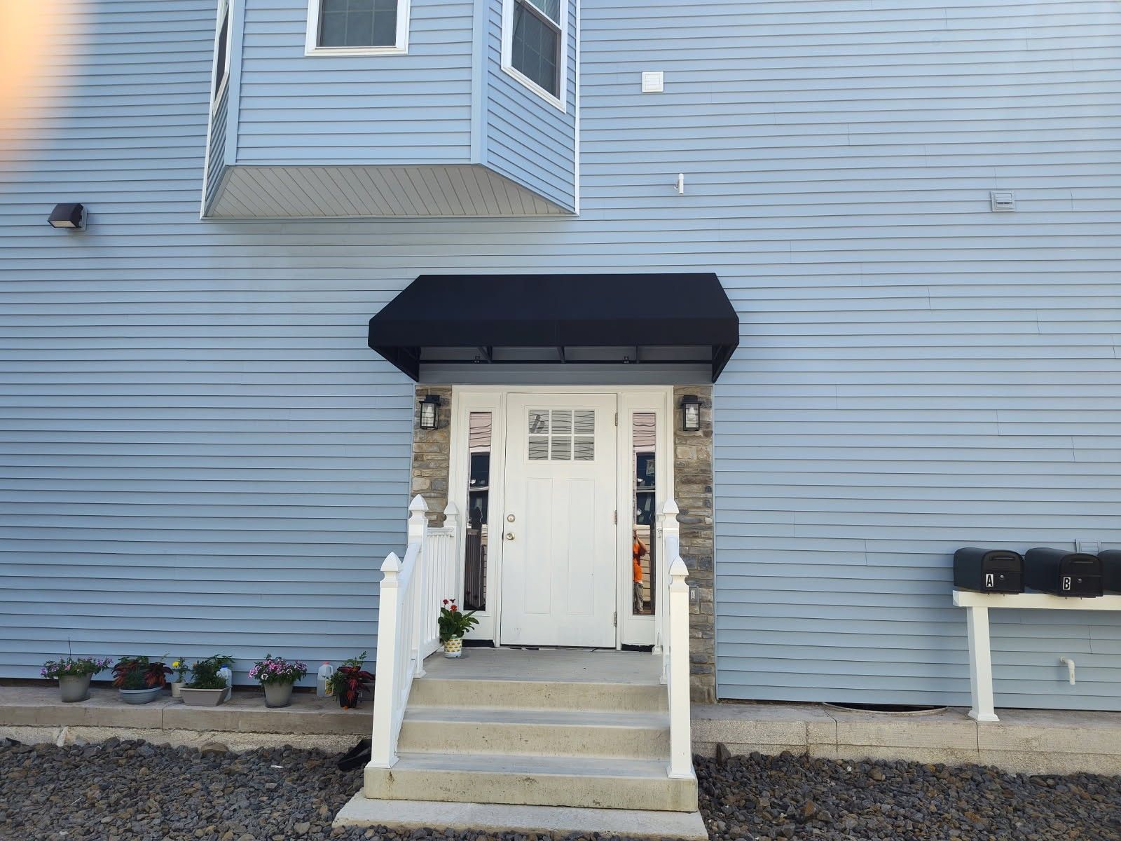A white front door with a black awning and short concrete stairs, set against light blue horizontal siding.