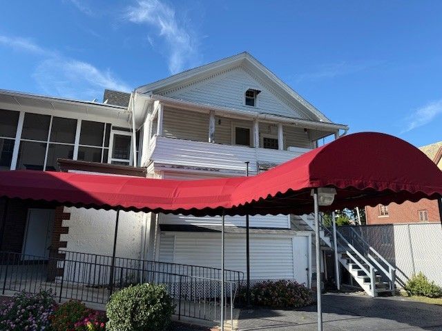 A white two-story house with a long red awning extending over a concrete driveway, metal railings, and an exterior staircase.