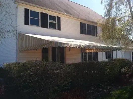 Two-story house with white siding, black shutters, and a long, striped fabric awning over the first-floor windows.