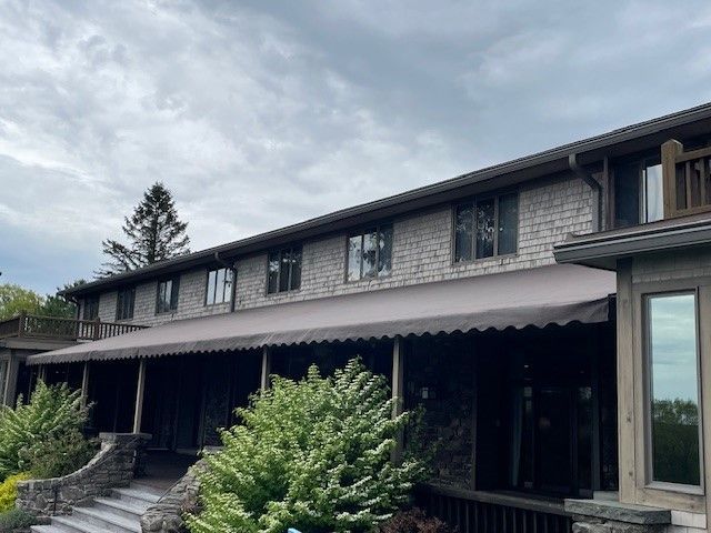 A two-story stone building with a long, gray fabric awning covering a shaded patio entrance under a cloudy sky.
