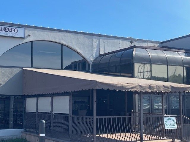 A white commercial building exterior featuring a covered outdoor patio area with a brown awning and a dark glass atrium.