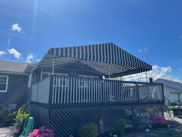 A dark gray house features a raised wooden deck with a black and white striped fabric awning under a bright blue sky.