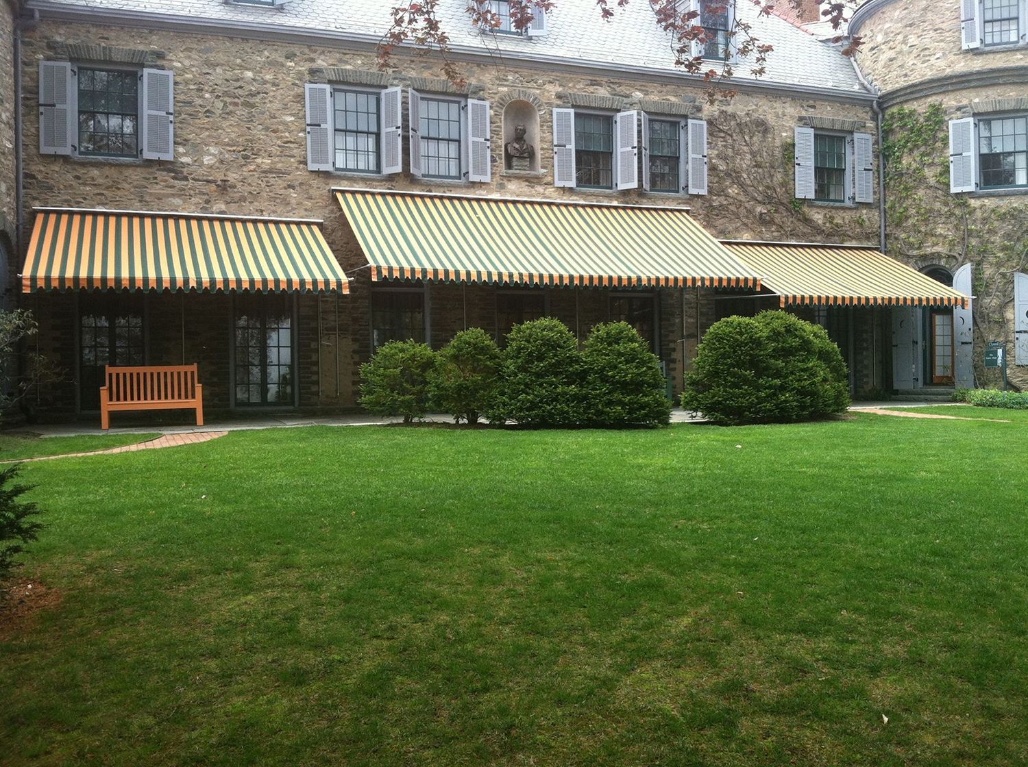 A stone house with three striped awnings, a wooden bench on the lawn, and round bushes in front of a patio area.