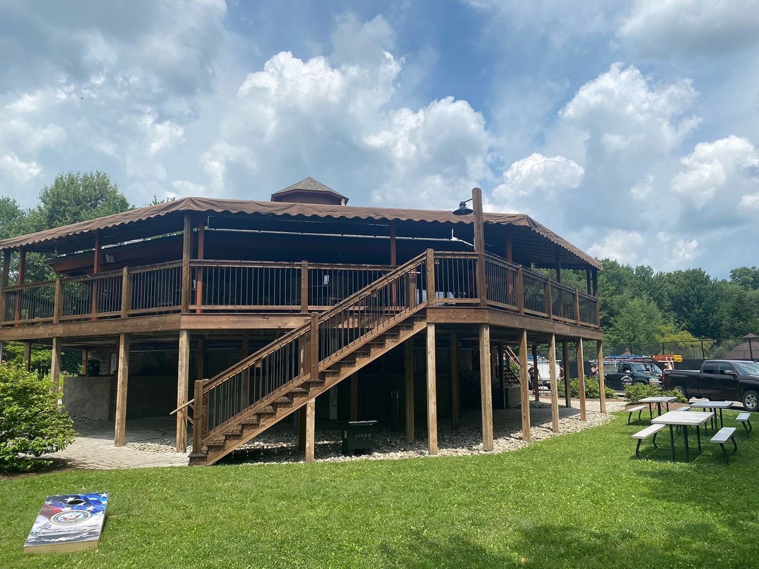 A large, raised, wooden gazebo structure with a staircase, surrounded by lawn and outdoor picnic tables.