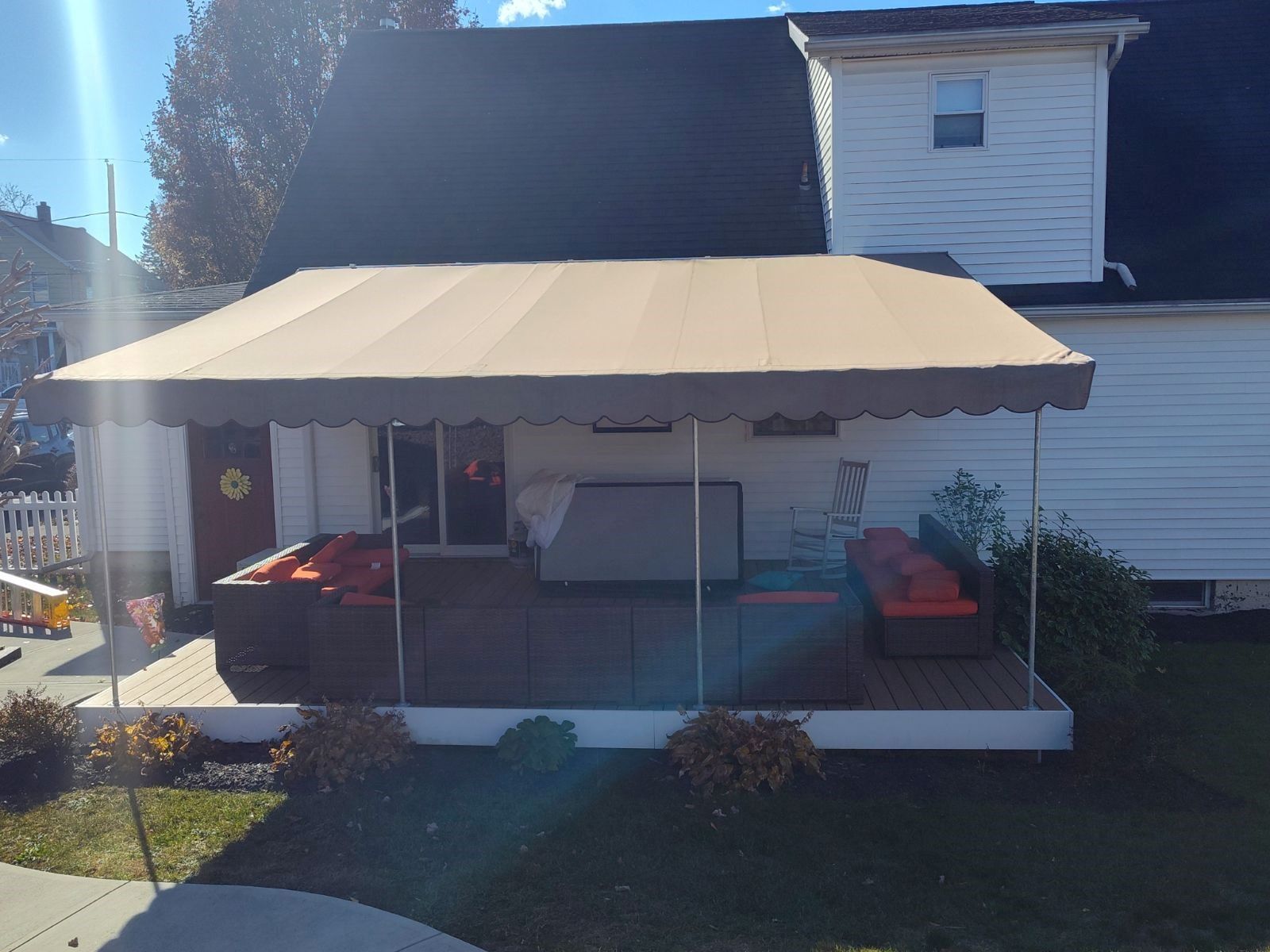 A tan patio awning covers a wooden deck with outdoor seating and a hot tub next to a white house.