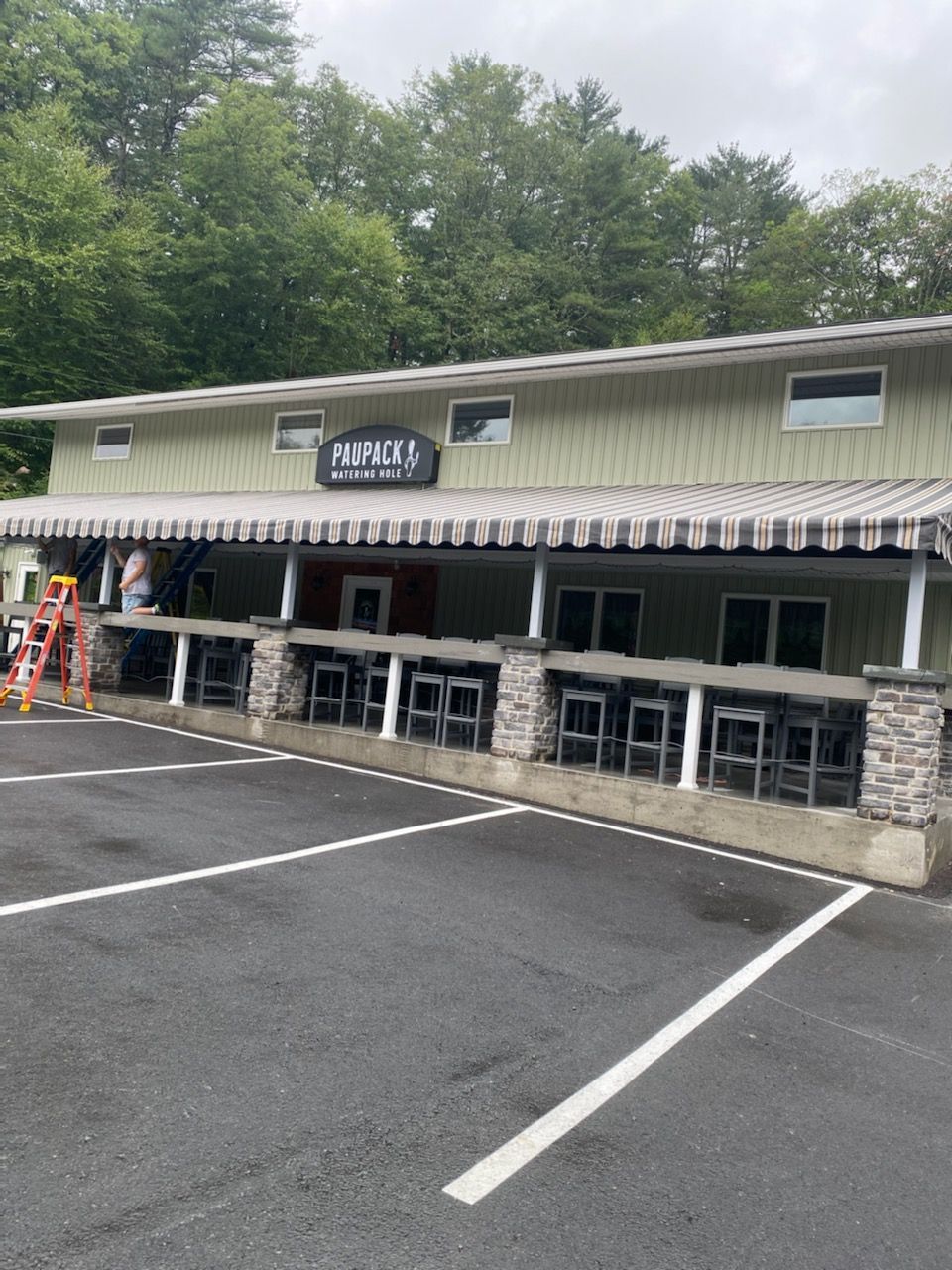 A light green building with a covered porch featuring stone pillars and bar seating, set in an asphalt parking lot.