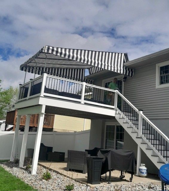 An elevated deck attached to a gray house, featuring a black-and-white striped awning and stairs leading to the patio below.