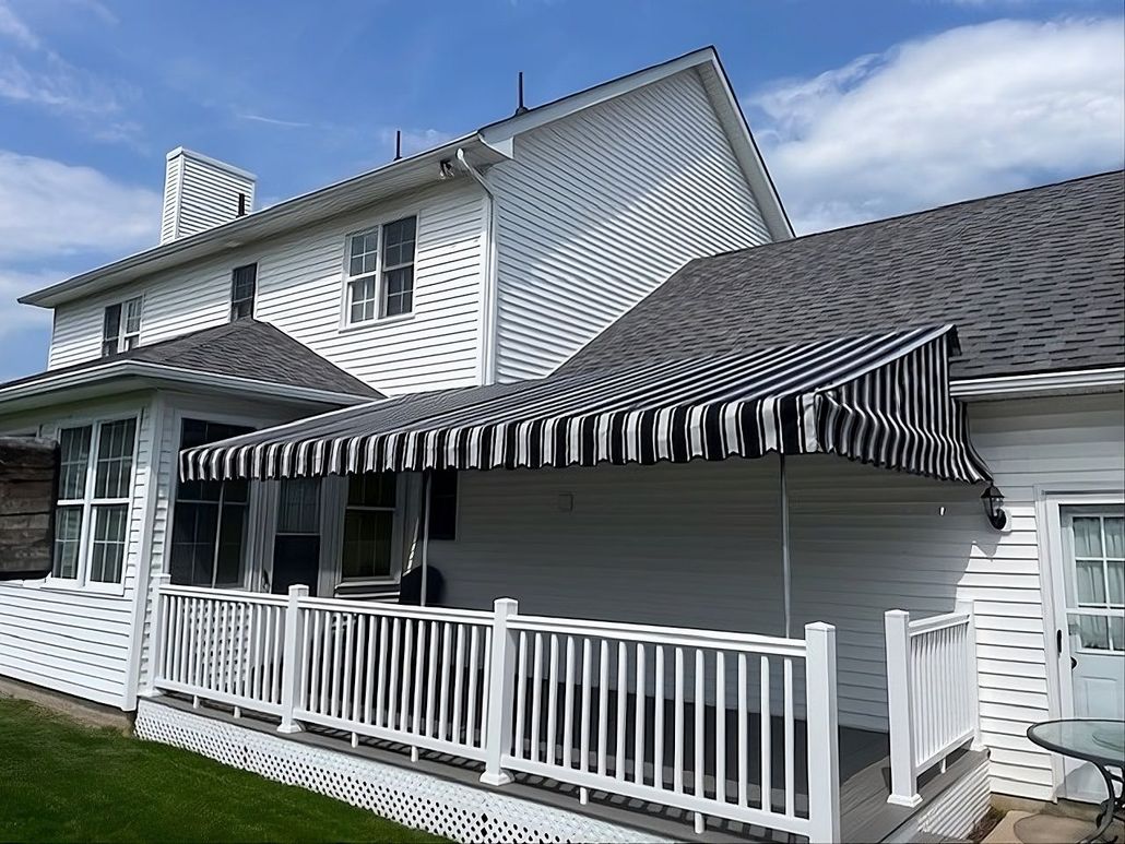 A white house with a deck featuring a black and white striped retractable awning under a blue sky.