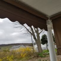 A brown retractable awning with a scalloped edge over a patio, looking out toward a house and field on a cloudy day.