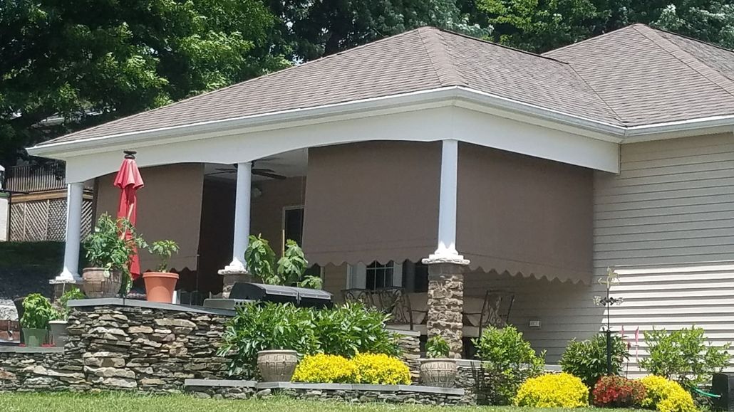 A covered patio with brown roll-down shades, stone retaining wall, potted plants, and landscaping against a beige house.