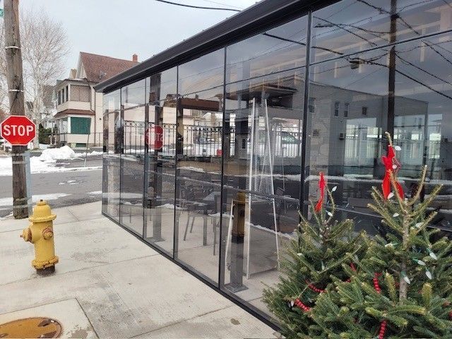 A glass-enclosed outdoor dining structure next to a sidewalk with a fire hydrant, stop sign, and small evergreen trees.