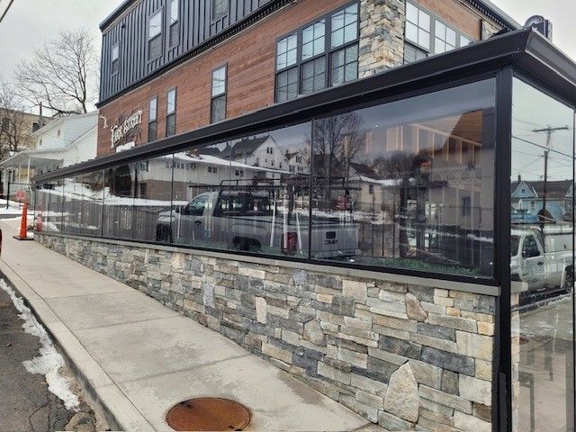 A sidewalk patio enclosed by a stone wall and glass panels, attached to a modern multi-story brick building.
