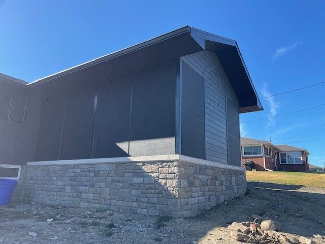 A residential house addition with grey horizontal siding, a stone base, and black-screened-in walls under a gable roof.
