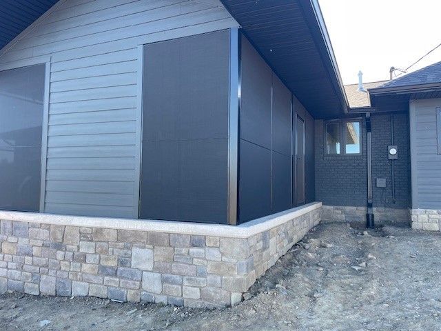 A house exterior featuring gray siding, stone veneer, and dark panels on a corner, with an unfinished dirt yard.