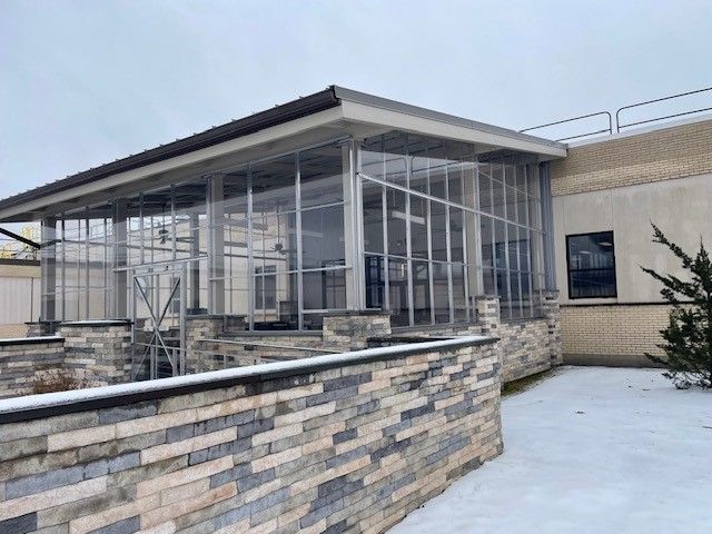 Exterior view of a building with a glass-enclosed structure. A stone wall runs along the ground in front. Snowy ground.