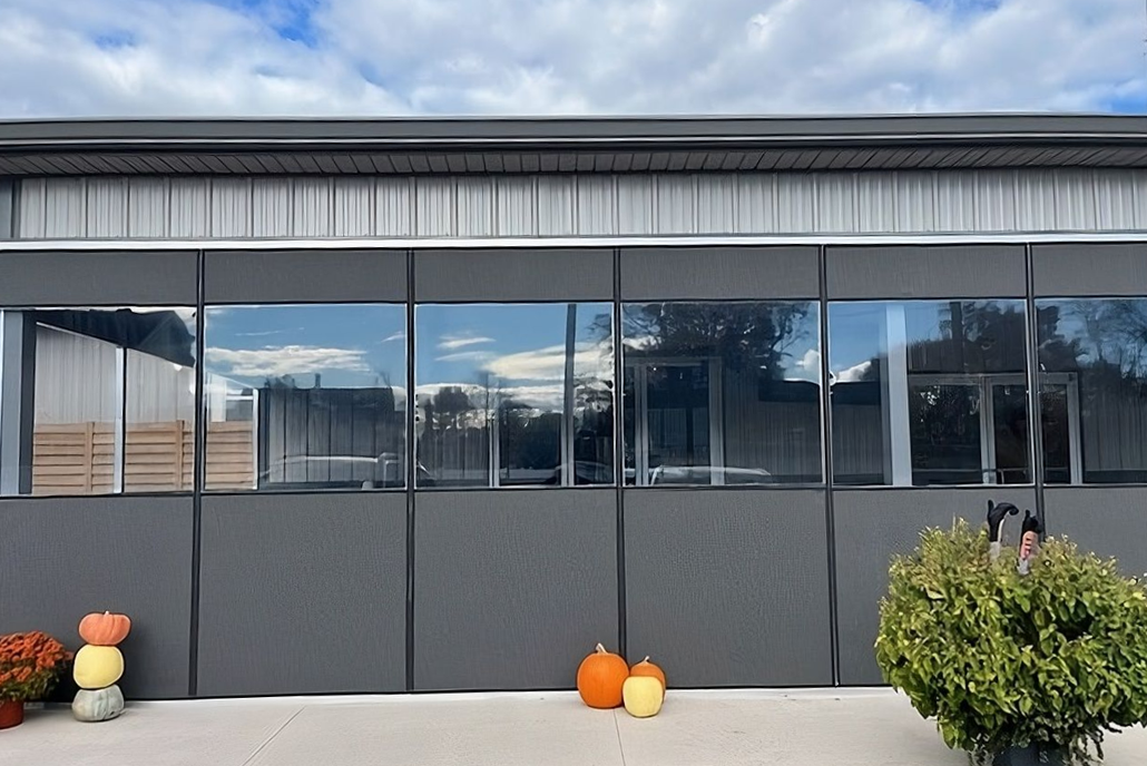 Gray building with large windows reflecting blue sky and trees, pumpkins and bush in the foreground.