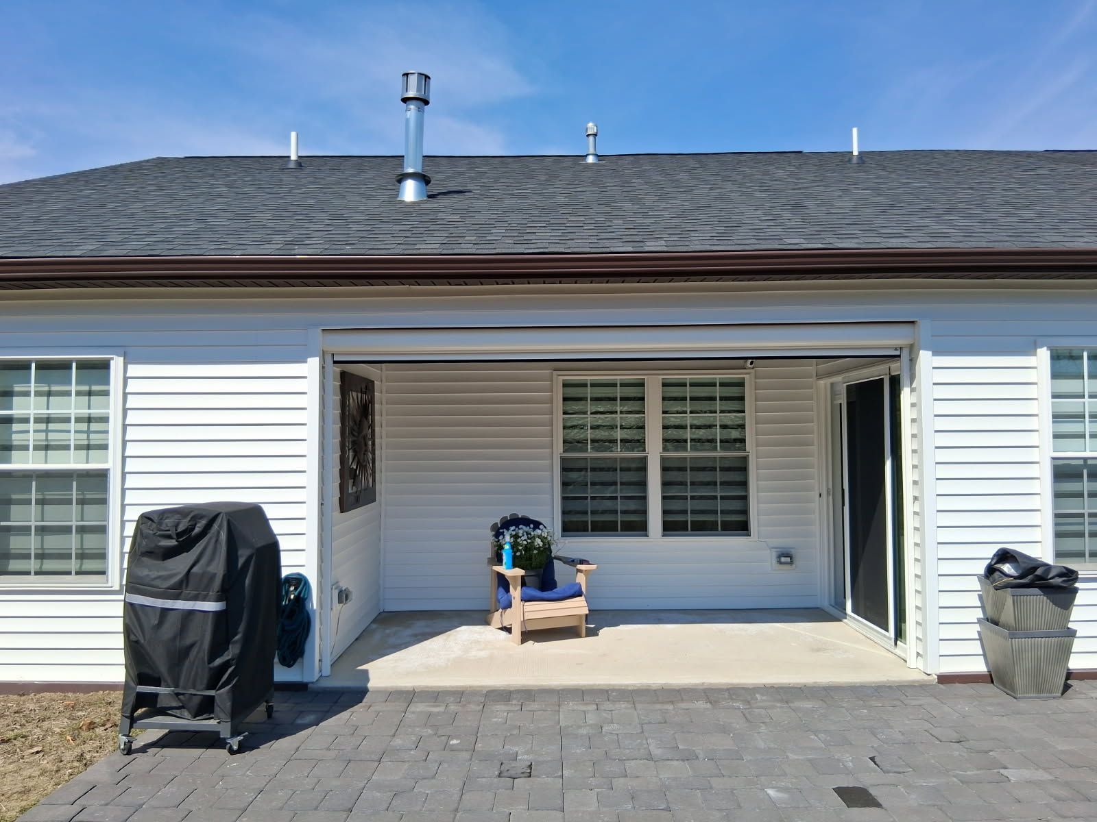 A covered patio with a wooden chair and a grill under a blue sky, attached to a house with white siding and gray shingles.