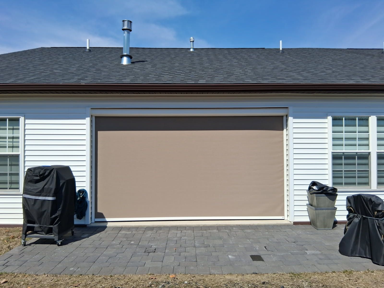 A beige retractable privacy screen pulled down over a patio doorway on the back of a house with two covered grills.