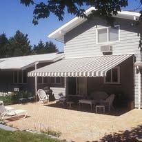 A patio with light-colored pavers, patio furniture, and a striped retractable awning attached to a gray house.