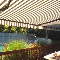 A brown-and-white striped awning extends over a wooden deck with green plants, a swimming pool, and a white fence nearby.