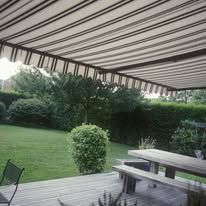 A striped awning shades a wooden deck with a picnic table and benches, overlooking a green backyard with shrubs.