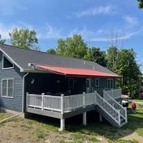 A gray house with a white deck, a set of stairs, and a large, extended red awning under a bright blue sky.